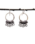 A pair of silver-toned dangle earrings with circular hoops and clusters of small dangling rings, hanging from a dark branch against a plain light background.