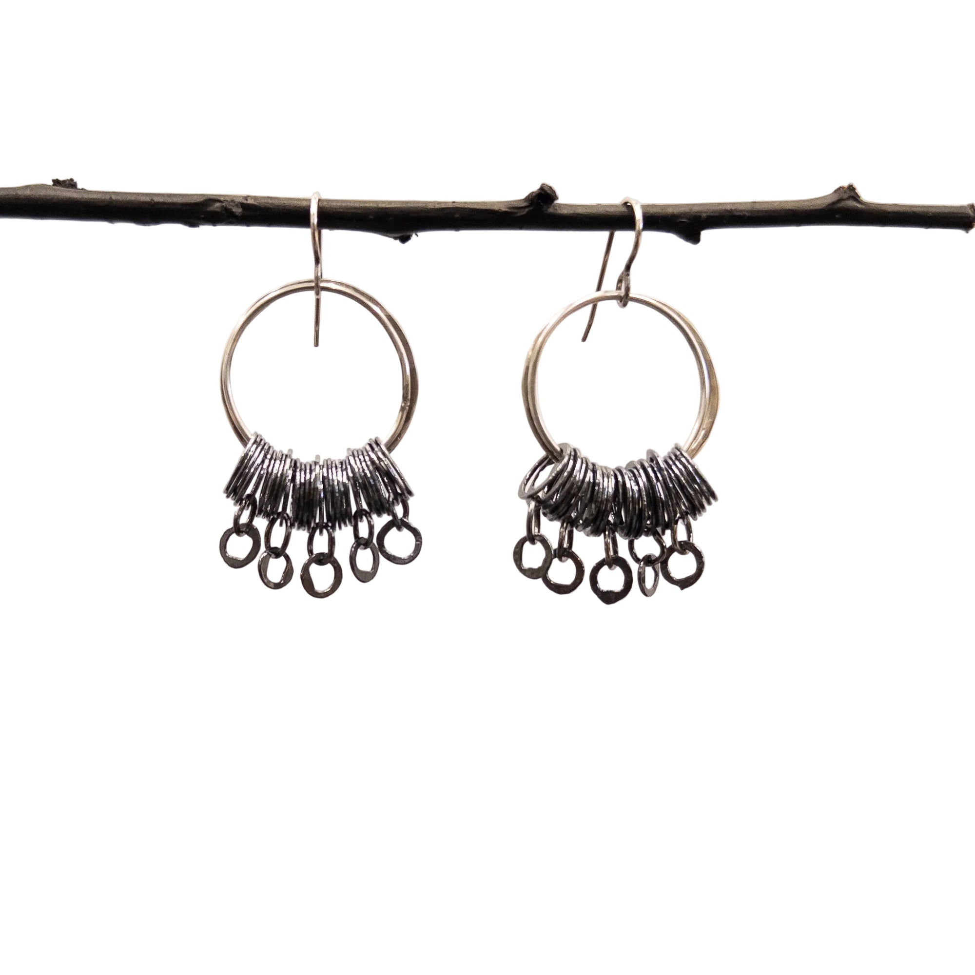 A pair of silver-toned dangle earrings with circular hoops and clusters of small dangling rings, hanging from a dark branch against a plain light background.